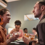 An image of a woman (left) stood talking and smiling with a man (right), as they both hold drinks.