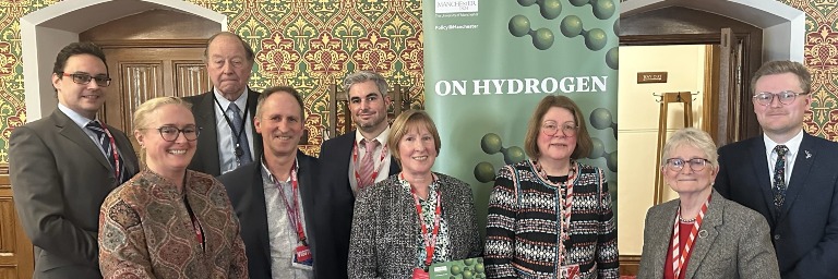 Nine academics and parliamentarians stand smiling in front of a green banner which reads "On Hydrogen"