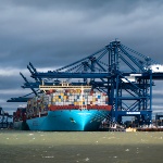 An image of a cargo ship docked underneath three blue cranes
