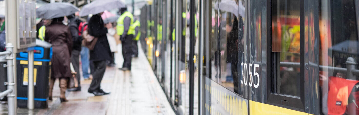 Image of a yellow Manchester tram waiting at the stop with people boarding, led by a conductor