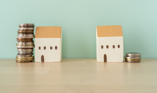 Two toy houses placed on a wooden table with coins stacked next to them. 